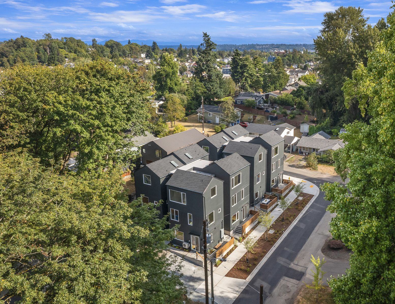 Drone view of the eight dark-grey gabled townhomes among trees, with the neighborhood beyond