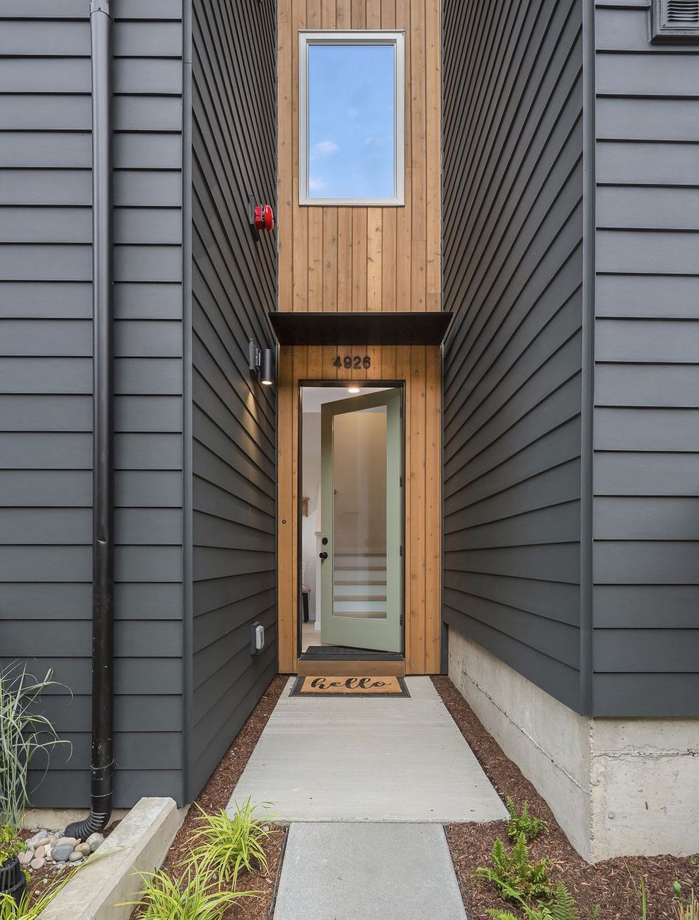 Narrow cedar-clad entry slot between two dark grey walls, sage green front door, hello mat