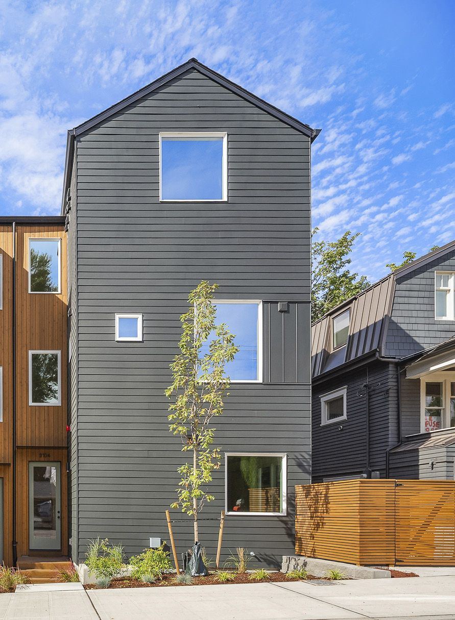 One townhome end-on: a tall dark-grey gable with a square window high up, a young tree, and a cedar fence
