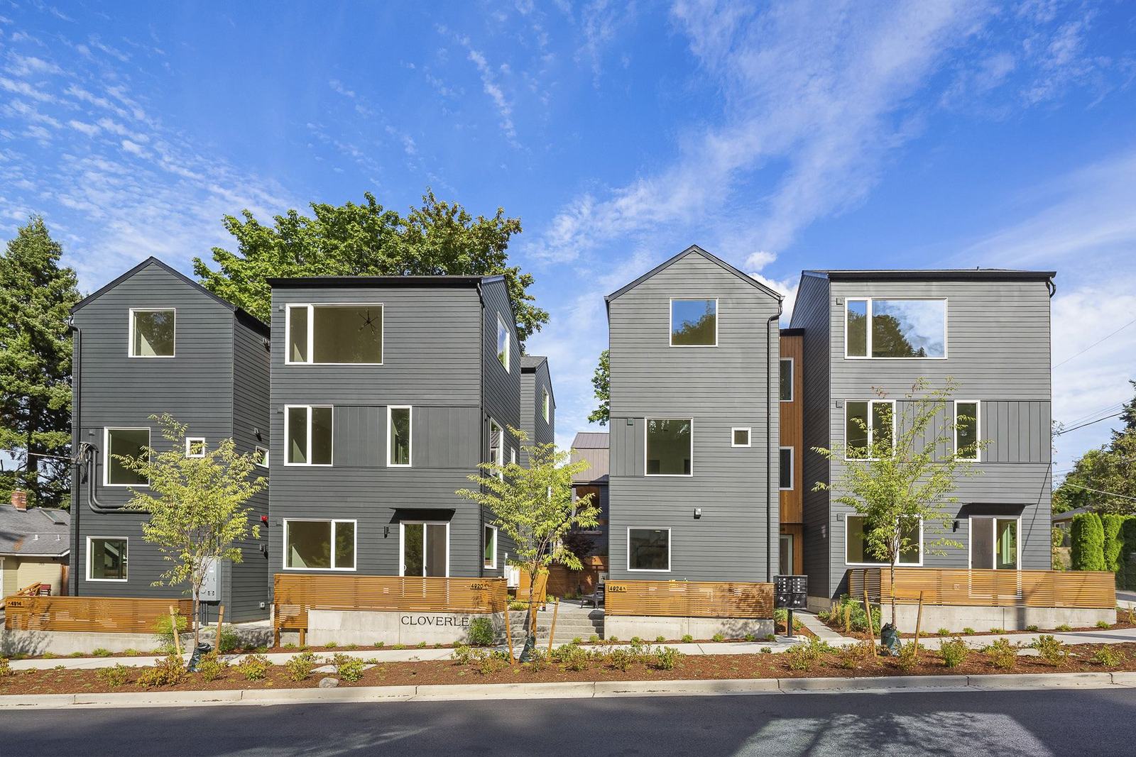 Four dark-grey gabled townhomes with cedar entry slots, seen from across the street under a blue sky