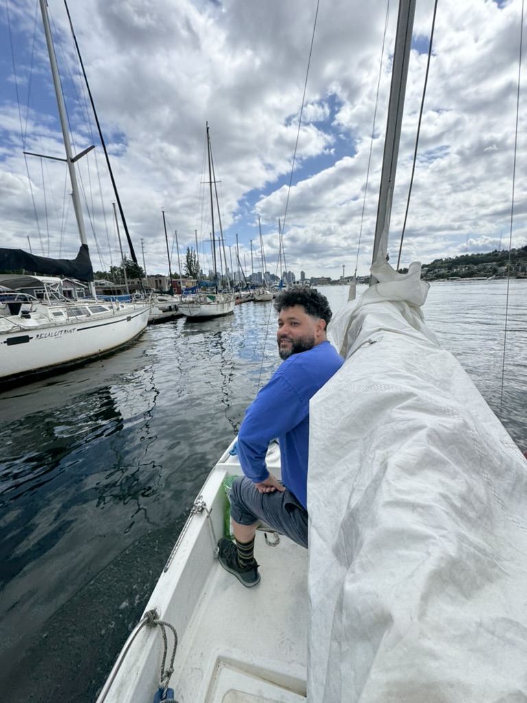 Biko Tabor sitting in Jasmine's cockpit beside the furled main, moorage and the Seattle skyline behind him.