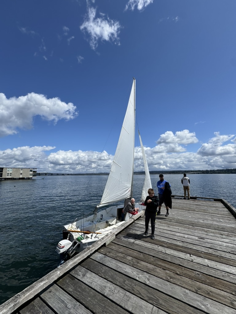 Jasmine alongside a wooden dock with sails up, three people on the dock and in the cockpit under a blue sky.