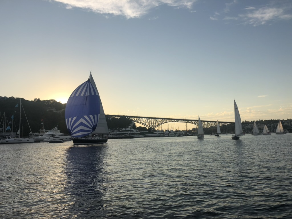 A blue and white spinnaker running downwind toward the Aurora Bridge in low evening light.