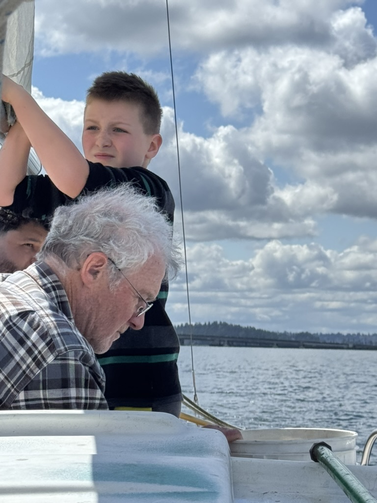 Wiley standing at the rigging looking to windward while B, white-haired and in a plaid shirt, works at the deck below, water and a floating bridge behind.