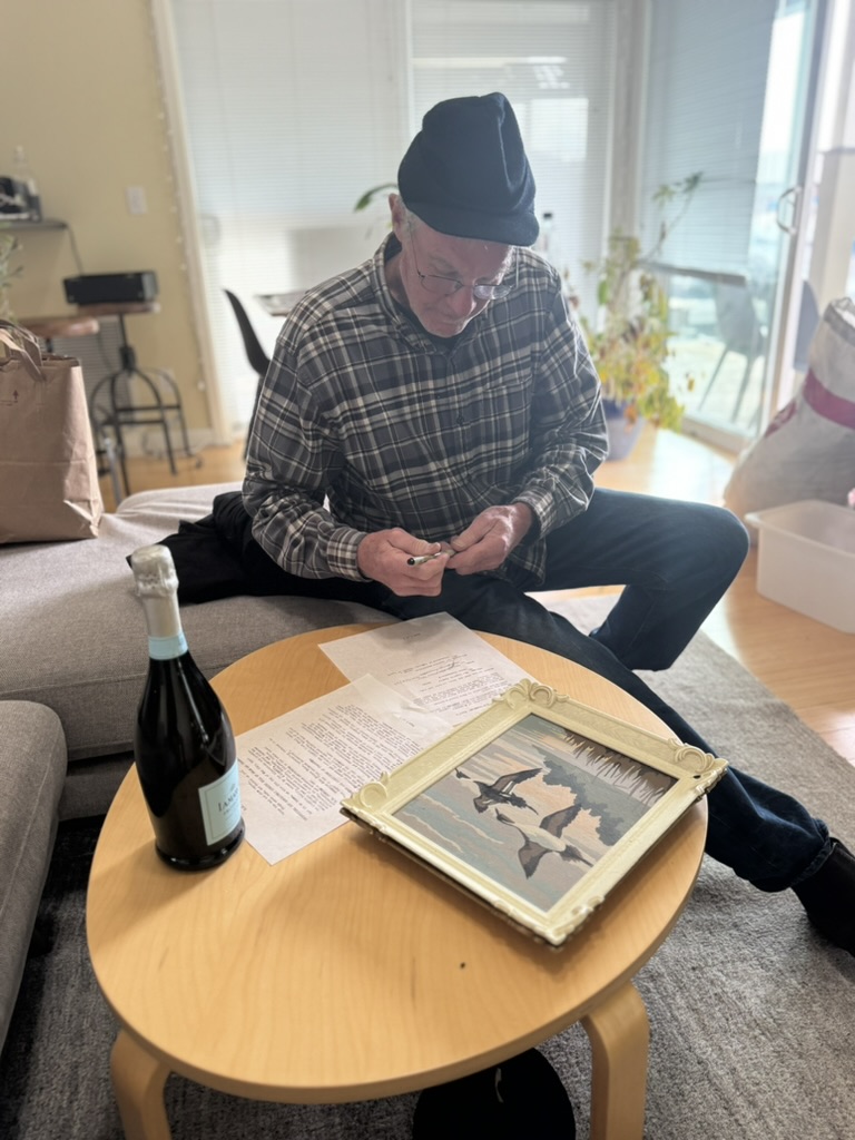 B signing the transfer document at a table, with a framed image of ducks and a bottle of champagne beside the papers.
