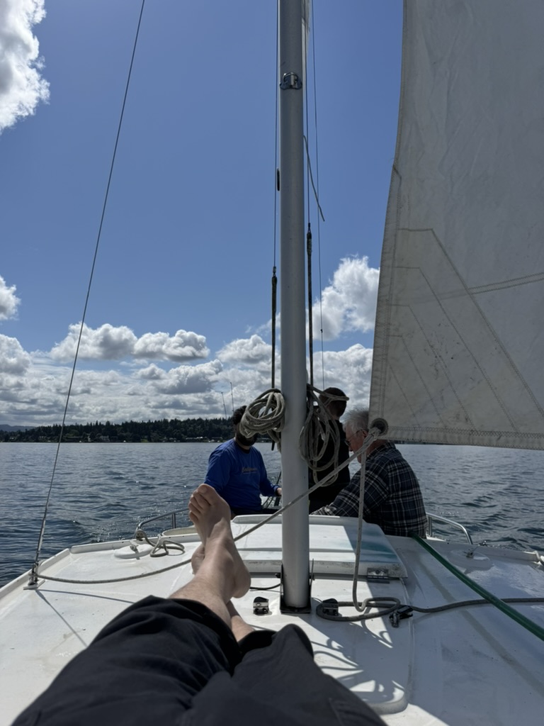 View forward from the cockpit under full sail, two crew at the helm, feet up in the foreground.